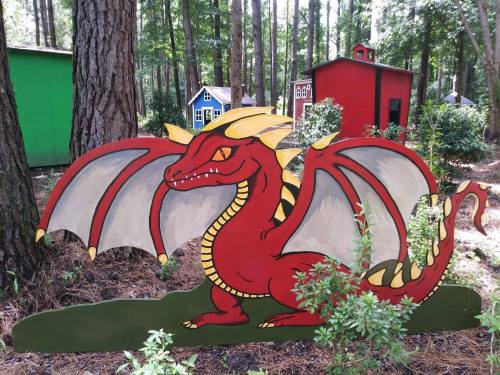 A colorful red dragon sculpture with large wings, set in a wooded area with small, colorful houses in the background.