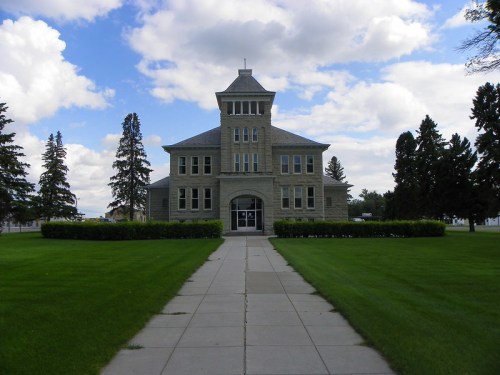 Historic stone building with a tower, surrounded by green grass and trees, under a partly cloudy sky.