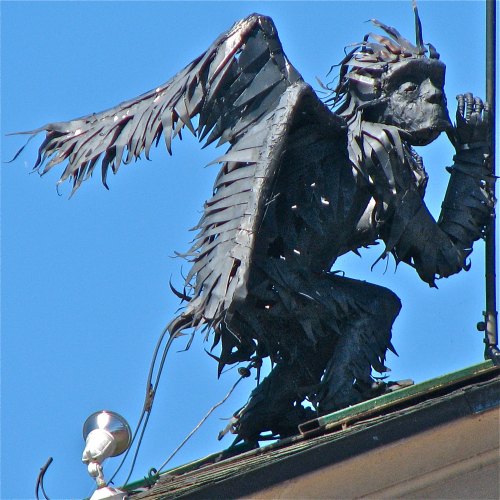 A metallic sculpture of a winged creature perched on a building, with intricate details and a blue sky background.
