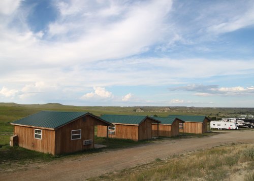 A row of wooden cabins with green roofs on a grassy landscape under a blue sky with scattered clouds.