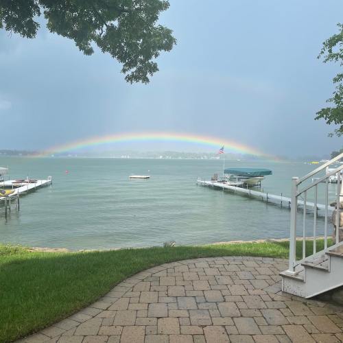 A serene lakeside view with a rainbow arching over the water, surrounded by greenery and boats docked nearby.