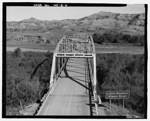 Black and white image of a bridge over the Little Missouri River, surrounded by rugged hills and scenic landscape.