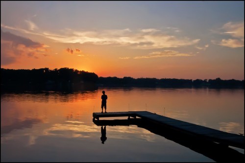 A person stands on a dock at sunset, reflecting on calm water with colorful clouds in the sky.