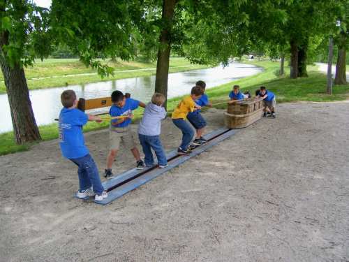 Children in blue shirts pull a wooden cart along a track in a park, with trees and a pond in the background.