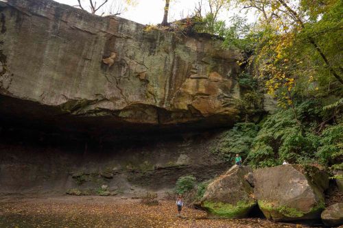 A rocky cliff with lush greenery above, two people exploring near large boulders by a calm, shallow water area.