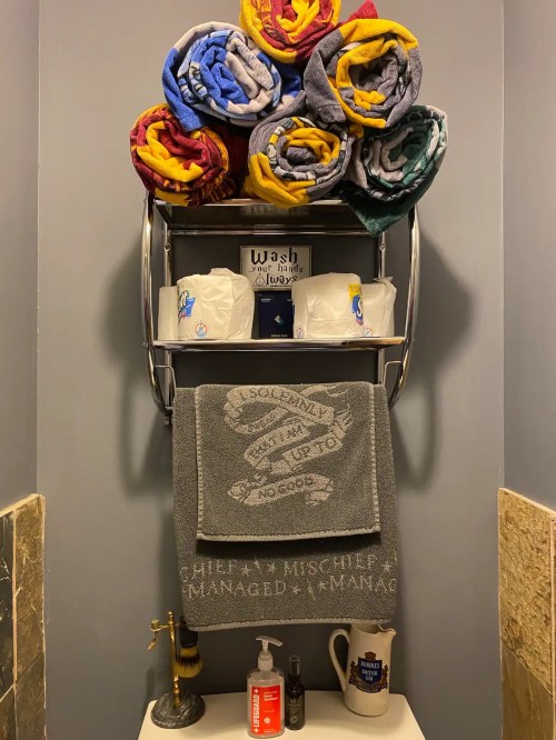 A towel rack with neatly stacked colorful towels, toilet paper, and a gray towel with decorative text on a bathroom wall.