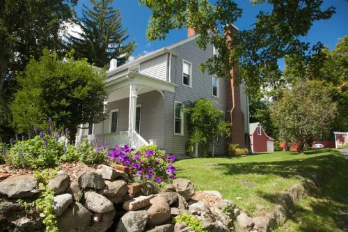 A charming gray house with a porch, surrounded by greenery and colorful flowers, set against a clear blue sky.
