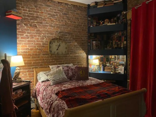 Cozy bedroom with a brick wall, a bed with patterned bedding, bookshelves, and warm lighting. Red curtains add color.