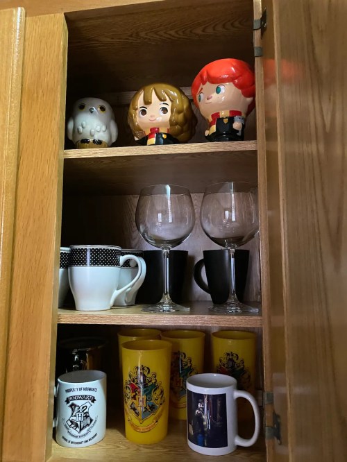 A wooden cabinet shelf displaying mugs, glasses, and figurines of characters, including an owl and two people.
