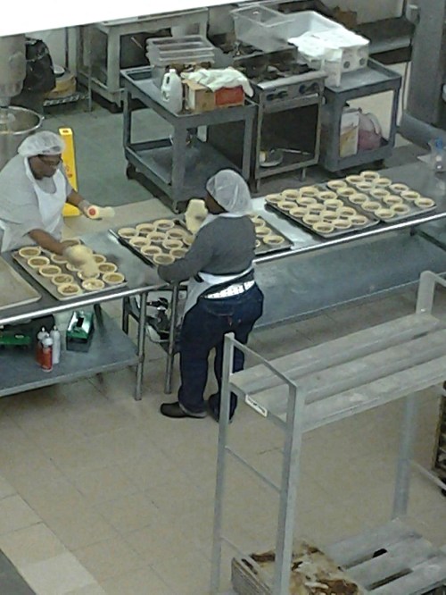 Two people in a kitchen preparing baked goods, with trays of dough on the counter and kitchen equipment in the background.