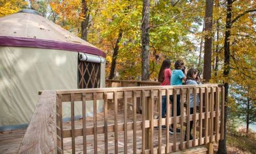 Three people stand on a wooden deck near a yurt, surrounded by colorful autumn trees.