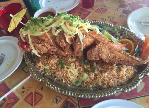 A large, fried fish garnished with onions and herbs, served on a bed of rice with colorful vegetables on a decorative platter.