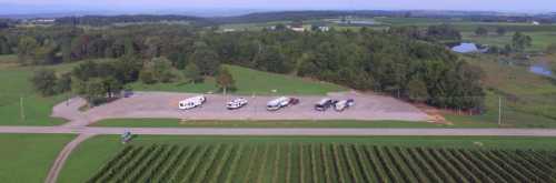 Aerial view of a parking area with several vehicles, surrounded by green fields and trees.