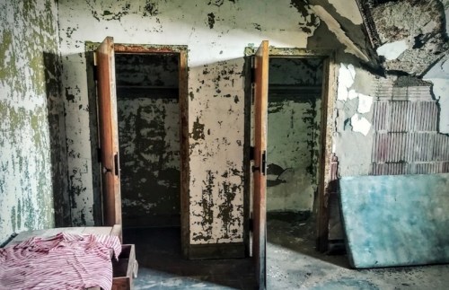 Abandoned room with peeling walls, two open doors, and a bed with a striped blanket.