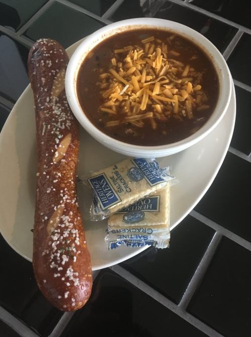 A plate with a soft pretzel, a bowl of chili topped with cheese, and two packets of crackers.