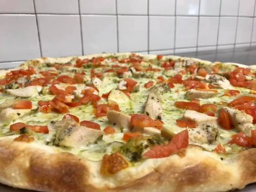 A close-up of a pizza topped with chicken, diced tomatoes, and green pesto on a golden crust.
