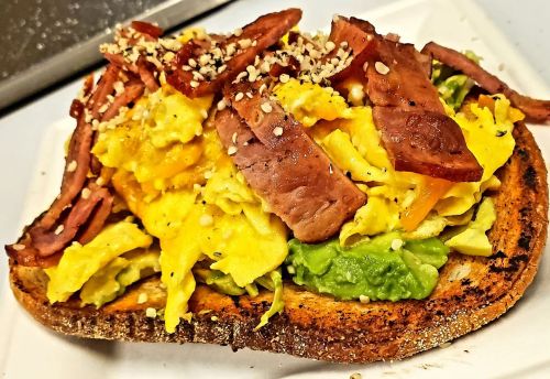 A hearty slice of toast topped with scrambled eggs, avocado, bacon, and sesame seeds.