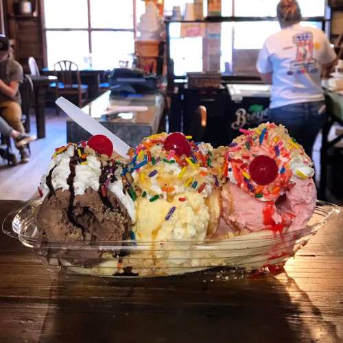 A colorful ice cream sundae with three scoops, topped with sprinkles, cherries, and chocolate sauce in a clear dish.