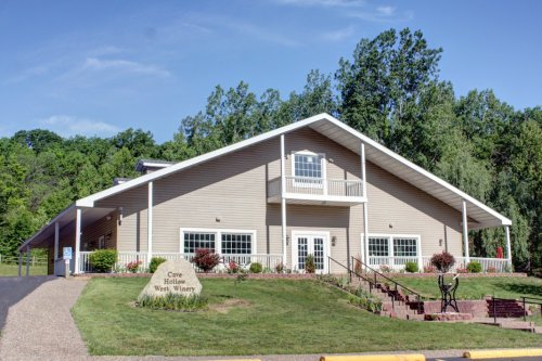 A large, modern winery building with a sloped roof, surrounded by greenery and a gravel pathway.
