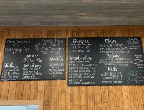 Menu board featuring burgers, sandwiches, plates, sides, and desserts with prices listed for each item.