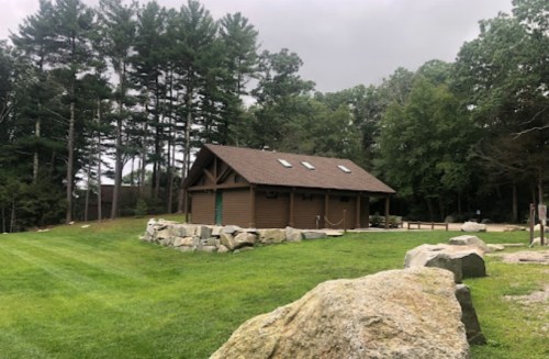 A brown building surrounded by trees and large rocks, set in a grassy area under a cloudy sky.