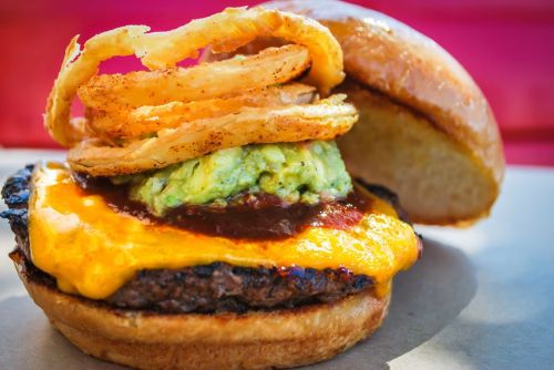 A close-up of a burger with melted cheese, guacamole, barbecue sauce, and crispy onion rings on a toasted bun.