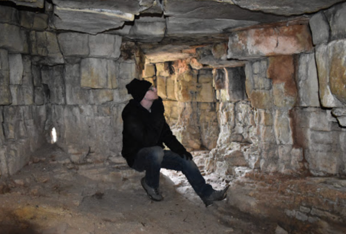 A person in a black coat and hat sits in a stone cave, surrounded by rough, ancient walls and dim lighting.