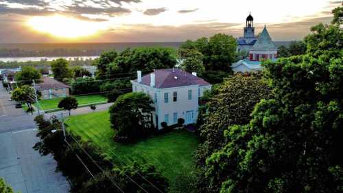 Aerial view of a historic house and a church at sunset, surrounded by lush greenery and a river in the background.