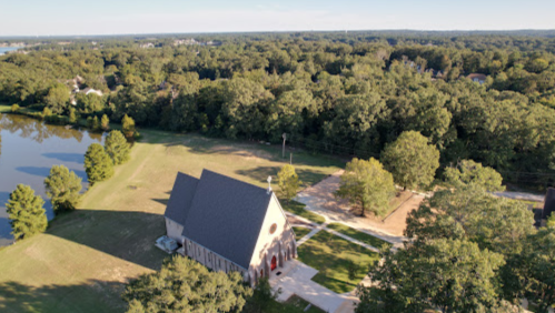 Aerial view of a church surrounded by trees and a pond, with a clear sky in the background.
