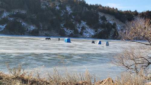 People ice fishing on a frozen lake, with blue tents set up against a backdrop of trees and a snowy hillside.