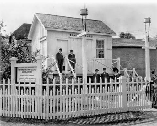 Historic black-and-white photo of a small house with a white picket fence and people standing outside.