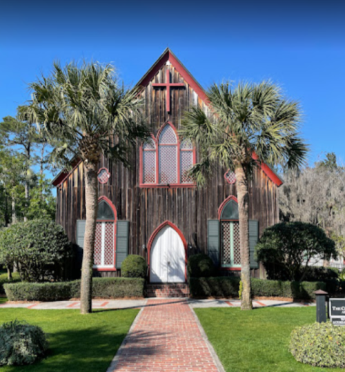 A charming wooden church with a red roof and cross, surrounded by palm trees and manicured greenery.