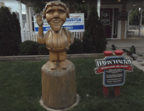 A wooden statue of a smiling man waving, next to a sign that reads "Wavin' Walton, Born May 22, 2009."