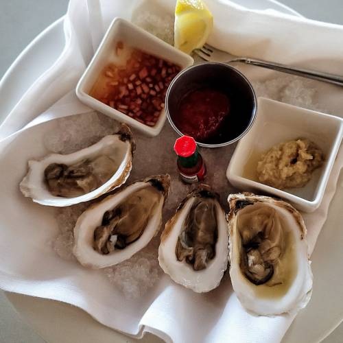 A plate of fresh oysters on ice, accompanied by lemon wedges and various dipping sauces.