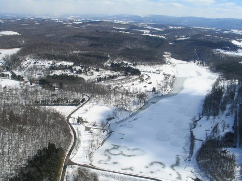 Aerial view of a snowy landscape with a frozen lake, winding roads, and scattered trees under a clear blue sky.