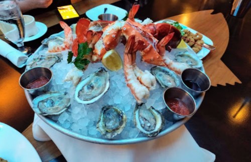 A seafood platter featuring oysters, shrimp, and lobster on ice, garnished with lemon and served with dipping sauces.