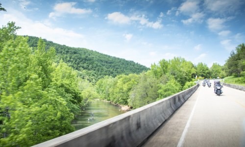A scenic road curves alongside a river, surrounded by lush green hills and trees, with motorcycles riding in the distance.