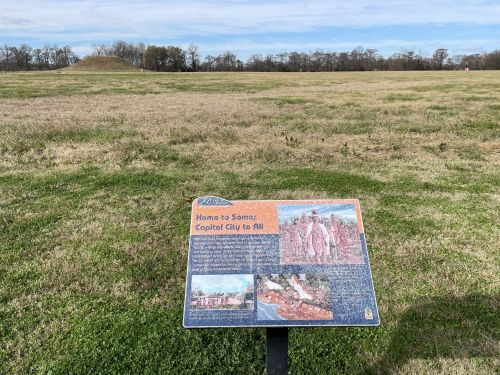 A sign in a grassy field, detailing the history of a site, with a mound visible in the background under a blue sky.
