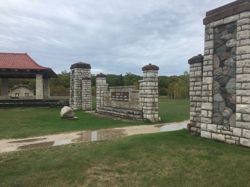 Stone entrance with pillars and a sign, leading to a grassy area with trees in the background under a cloudy sky.