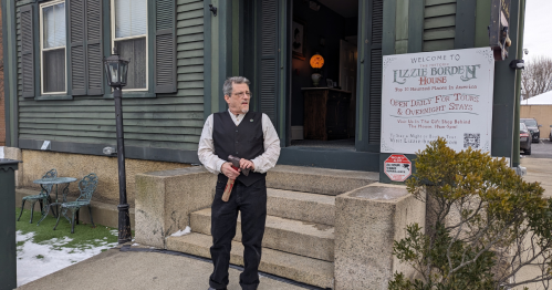 A man in period clothing stands outside the Lizzie Borden House, with a sign welcoming visitors. Snow is on the ground.