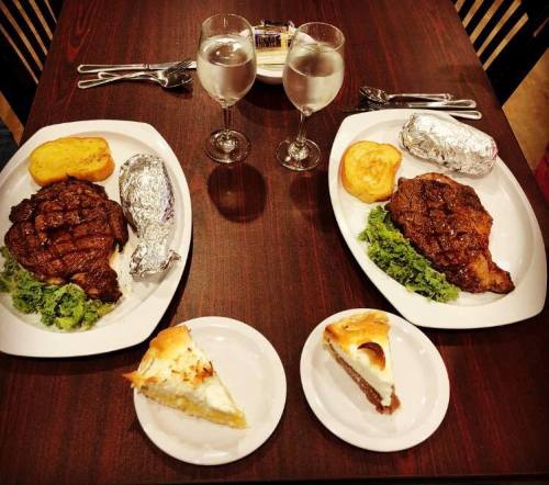 Two plates of steak with sides of baked potatoes, cornbread, and salads, plus two desserts on a table with water glasses.