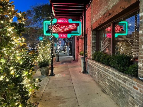 A charming street scene at dusk featuring a neon sign for "Weidmann's" and festive lights along the sidewalk.