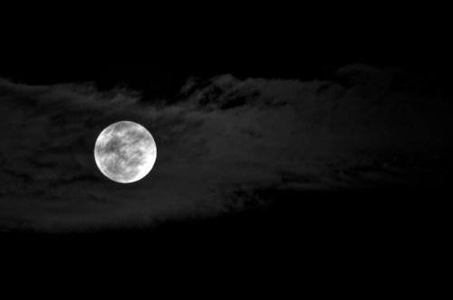 A full moon illuminated against a dark sky, partially obscured by wispy clouds.