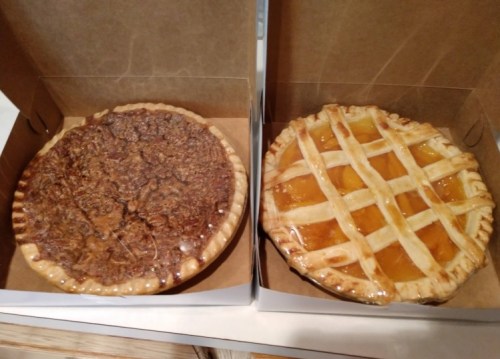 Two pies in cardboard boxes: one pecan pie on the left and one peach pie with a lattice crust on the right.