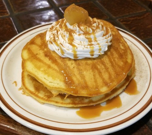 Two fluffy pancakes topped with whipped cream, caramel drizzle, and a slice of fruit on a white plate.