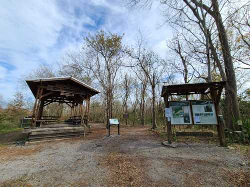 A wooden pavilion and information boards in a wooded area with bare trees under a cloudy sky.