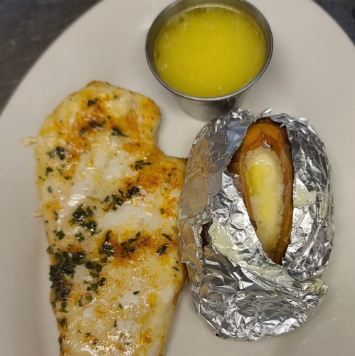 Grilled fish fillet with herbs, a baked potato wrapped in foil, and a small cup of melted butter on a plate.