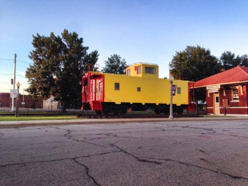 A bright yellow and red train caboose parked beside a building on a quiet street, with trees in the background.