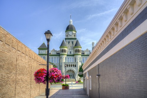 Historic building with a clock tower, surrounded by colorful flowers, viewed from a narrow pathway. Clear blue sky above.
