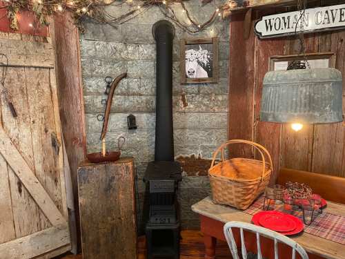 Cozy rustic interior with a wood stove, wooden decor, a basket, and a table set for dining. Warm, inviting atmosphere.